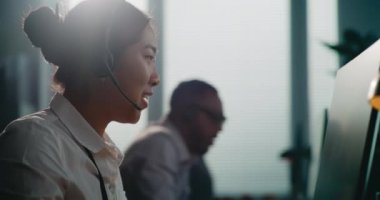 Asyalı yardım hattının operatörünün kulaklığı takılı vaziyette telefonda müşteriye çevrimiçi yardım sağlarken görüntüsünü kapatın. Modern çağrı merkezinde çeşitli teknik müşteri destek uzmanları çalışıyor..