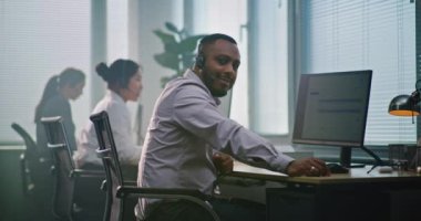 Kulaklıklı Afro-Amerikan teknik destek operatörü, bilgisayarın önünde oturuyor, müşteri hizmetleri sunan kameraya bakıyor. Çeşitli yardım masası uzmanları modern çağrı merkezinde çalışıyor..