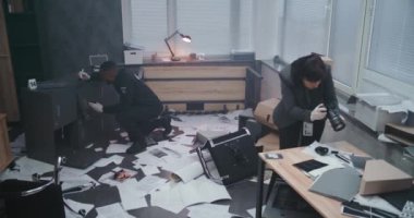 Ofis soygunu davası: Kadın adli tıp uzmanı ipuçlarının ve kanıtların fotoğraflarını çekiyor, Afro-Amerikan polisi parmak izlerini araştırıyor. Takım elbiseli erkek dedektifin olay yerini incelemesi..