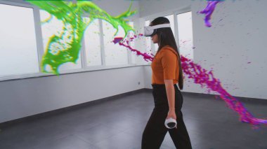 Sanal gerçeklikteki Asyalı genç kadın kablosuz kontrolörlerle resim yapıyor, soyut çevrimiçi sanat eserleri yaratmak için jestler yapıyor. Boya damlalarının holografik 3 boyutlu animasyonu. Gelişmiş gerçeklik teknolojisi.