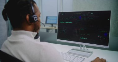 Afrika kökenli Amerikalı profesyonel finans uzmanı. Kulaklık analizi. Hisse senetleri ve hisse senetleri. PC ekranında grafikler ve diyagramlar var. Borsa şirketinin modern ofisi. E-iş.