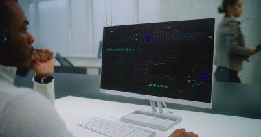 Afrikalı Amerikalı adam, uluslararası şirketin modern ofisinde çalışıyor: kulaklık takan borsacı, hisse senetlerini bilgisayar, grafik ve diyagram kullanarak izliyor. E-iş.