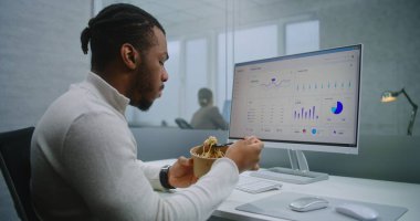 Afro-Amerikalı finans uzmanı ya da E-ticaret müdürü öğle arasında iş yerinde yiyecek yiyor. PC ekranında ticari veriler ve diyagramlar var. E-ticaret şirketinin modern ofisi.