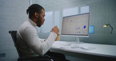 Afro-Amerikalı finans uzmanı ya da E-ticaret müdürü öğle arasında iş yerinde yiyecek yiyor. PC ekranında ticari veriler ve diyagramlar var. E-ticaret şirketinin modern ofisi.