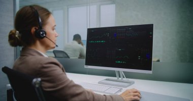Profesyonel kadın borsacı kulaklıkla konuşur, finansal tabloları ve bilgisayardaki şemaları analiz eder. Bilgisayar ekranında borsa ve hisse senetleri görülüyor. Borsa şirketinin modern ofisi. E-ticaret.