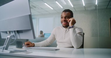 Heyecanlı genç işadamı müşteriyle başarılı iş anlaşmasını kutluyor, bilgisayar kullanıyor, cep telefonuyla konuşuyor. Afrika kökenli Amerikalı pazarlama uzmanı modern ofiste çalışıyor. E-ticaret