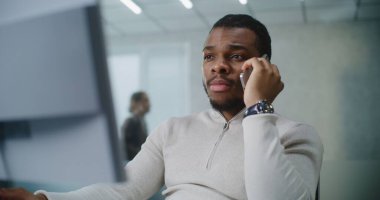 Afrikalı Amerikalı adam parlak gelişmiş bir ofiste çalışıyor: Genç pazarlama uzmanı veya müşteri yöneticisi iş ortağıyla konuşuyor veya cep telefonuyla müşteriye danışıyor, bilgisayar kullanıyor. E-iş konsepti.