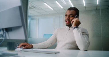 Genç Afrikalı Amerikalı işadamı iş ortağıyla anlaşma hakkında konuşuyor, cep telefonundan müşterilerine danışıyor, bilgisayar kullanarak verileri izliyor. Pazarlama uzmanı parlak modern ofiste çalışıyor. E-ticaret.