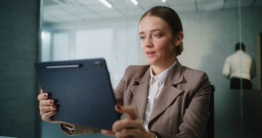 Beyaz iş kadını iş yerinde oturup tablet bilgisayar kullanarak iş ortağı veya müşteriyle görüntülü konuşma yapıyor. Modern ofiste pazarlama müdürü veya finans uzmanı olarak çalışıyor. E-ticaret.