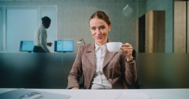 Modern ofis: İş yerinde oturan, kahve içen, gülümseyen ve kameraya bakan başarılı iş kadını veya pazarlama profesyonelinin portresi. Afrikalı Amerikalı meslektaşım arka planda çalışıyor.