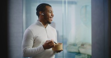 Afro-Amerikan ofis çalışanı ya da işadamı yemek yiyor, iş stratejisini ortağıyla kablosuz kulaklık kullanarak tartışıyor. E-ticaret şirketinin modern ofisinde pazarlama uzmanı.