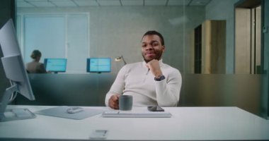 Modern ofis: Genç Afrikalı Amerikalı iş adamının portresi ya da iş yerinde elinde fincanla oturan, gülümseyen ve kameraya bakan başarılı pazarlama uzmanı. Bayan meslektaşım arka planda çalışıyor..