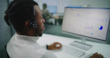 Afrikalı Amerikalı pazarlama uzmanı müşteriyle kulaklıkla konuşuyor, bilgisayarda çalışıyor, çevrimiçi müşteri hizmetleri sağlıyor. PC ekranında ticari veriler ve grafikler var. E-ticaret şirketinin modern ofisi