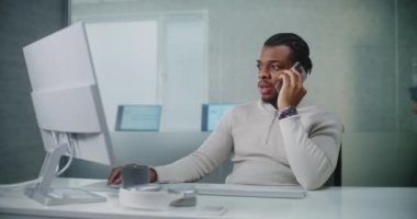Genç Afrikalı Amerikalı işadamı iş ortağıyla anlaşma hakkında konuşuyor, cep telefonundan müşterilerine danışıyor, bilgisayar kullanarak verileri izliyor. Pazarlama uzmanı parlak modern ofiste çalışıyor. E-ticaret.