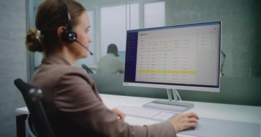 Beyaz kadın E-ticaret şirketinin modern ofisinde çalışıyor: Müşteri ile kulaklık iletişiminde kadın müşteri yöneticisi veya finans uzmanı, ticari veri ve grafikleri bilgisayar kullanarak analiz ediyor.