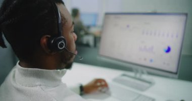 Profesyonel Afro-Amerikan finans uzmanı ya da elektronik ticaret yöneticisi, kulaklık danışmanlığı müşterisi, bilgisayar üzerinde çalışıyor. Ticari verileri ve diyagramları olan bilgisayar ekranı. E-ticaret şirketinin modern ofisi