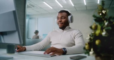Kulaklık takan Afrikalı Amerikalı adam modern ofiste Noel süslemeleri ile çalışıyor: Başarılı pazarlama uzmanı veya müşteri yöneticisi müzik dinliyor, bilgisayarda çalışıyor. E-iş konsepti.