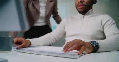 Afrika kökenli Amerikalı pazarlama uzmanı ya da elektronik ticaret yöneticisi bilgisayar kullanıyor ve klavyeye veri giriyor. Bayan yönetici meslektaşına yardım ediyor. Uluslararası E-ticaret şirketinin modern ofisi.