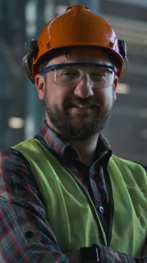 Üretim fabrikasında koruyucu üniforma, kask ve gözlük takan profesyonel ağır endüstri çalışanı. Erkek teknisyen ya da mühendis gülümser ve kameraya bakar. Dikey çekim