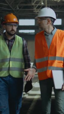 Güvenlik üniformalı iki ağır endüstri mühendisi ve sert şapkalı iki kişi üretim tesisinde yürüme sürecini tartışıyor. Olgun yönetici dizüstü bilgisayarı tutuyor, fabrikadaki yetişkin işçilerle konuşuyor. Dikey çekim