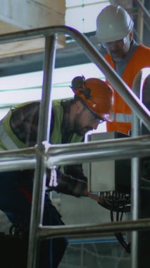 Üretim tesisinde veya fabrikada koruyucu üniforma ve kask giyen iki profesyonel ağır endüstri mühendisi. Erkek işçi konuşur ve yetişkin yöneticilere makine ekipmanları gösterir. Dikey çekim