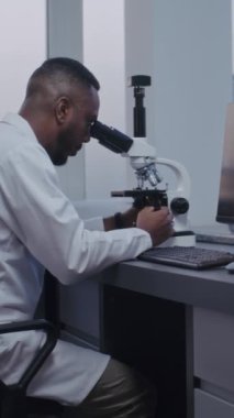 Dikey çekim yapan Afrikalı Amerikalı mikrobiyolog mikroskop altında test örneklerini yüksek teknoloji kullanarak analiz ediyor. Bilgisayar ekranında hücrelerin veya bakterilerin görüntüsü var. Mikrobiyoloji.