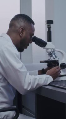 Dikey çekim yapan Afrikalı Amerikalı mikrobiyolog mikroskop altında test örneklerini yüksek teknoloji kullanarak analiz ediyor. Bilgisayar ekranında hücrelerin veya bakterilerin görüntüsü var. Mikrobiyoloji.