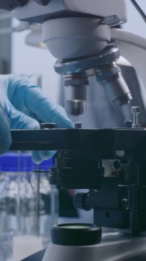 Profesyonel mikrobiyolog ya da eldivenli bilim adamının dikey görüntüsü analiz etmek ve ilaç geliştirmek için test numunesiyle birlikte cam kaydırağı mikroskop altına koyuyor. Yüksek teknoloji laboratuvar aygıtı.