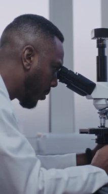 Dikey çekim yapan Afrikalı Amerikalı mikrobiyolog mikroskop altında test örneklerini yüksek teknoloji kullanarak analiz ediyor. Bilgisayar ekranında hücrelerin veya bakterilerin görüntüsü var. Mikrobiyoloji.