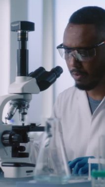 Beyaz önlüklü bilim adamları ileri laboratuarda çalışıyorlar. Afrikalı Amerikalı mikrobiyolog test tüplerindeki sıvıyı mikroskop altında inceliyor. Dikey çekim