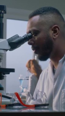 Beyaz önlüklü Afrikalı Amerikalı erkek mikrobiyolog veya kimyager aşı geliştirmek için mikroskop altında yeni virüs örneklerini inceliyor. Arka planda çalışan kadın bilim adamı. Dikey çekim