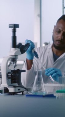 Tıp laboratuvarı: Afrikalı Amerikalı mikrobiyolog test tüpünden sıvı alır, mikroskop altında inceler, yüksek teknolojili ekipman kullanarak örnek analizi yapar. Dikey çekim