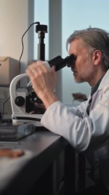 Olgun bilim adamı ya da mikrobiyolog mikroskop altında fosil analizi yapar, dizüstü bilgisayar kullanır. Eski insan iskeletinin kemikleri ve masada duran aletler. Dikey çekim