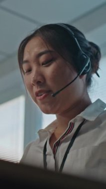 Dikey çekim, Asyalı kadın teknik müşteri destek uzmanının düşük açılı portresi kulaklıkla konuşuyor, bilgisayarda çalışıyor, müşterilere çevrimiçi yardım sağlıyor. Yardım hattı operatörü çalışıyor.