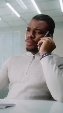 Genç Afrikalı Amerikalı işadamı iş ortağıyla anlaşma hakkında konuşuyor, cep telefonundan müşterilerine danışıyor, bilgisayar kullanarak verileri izliyor. Pazarlama uzmanı parlak modern ofiste çalışıyor. Dikey