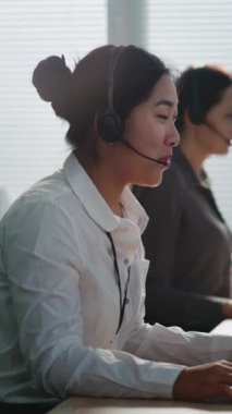 Teknik müşteri destek uzmanlarından oluşan bir ekibin dikey görüntüsü. Asyalı kadın yardım hattı operatörü kulaklıkla konuşuyor, bilgisayarda çalışıyor, müşterilere çevrimiçi yardım sağlıyor.