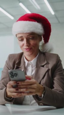 Noel Baba şapkalı kadın pazarlama uzmanı ya da E-ticaret müdürü iş yerinde oturuyor, cep telefonu kullanarak kurye uygulamasında iş ortağıyla sohbet ediyor. İş kadını modern ofiste çalışıyor. E-iş.