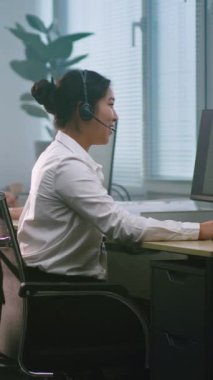 Modern çağrı merkezinde çalışan çok ırklı teknik destek operatörleri ekibi. Asyalı kadın danışma uzmanı kulaklıkla konuşuyor, müşteri için çevrimiçi teknik yardım sağlarken bilgisayar kullanıyor