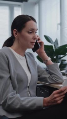 Kadın finans analisti, iş arkadaşıyla cep telefonu kullanarak proje stratejisini tartışıyor. Pazarlama bölümünden uzman, PC monitöründe gösterilen parametrelerle belgelerdeki verileri kontrol ediyor.