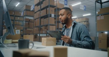 Afro-Amerikan depo çalışanı paketleri tablet bilgisayar kullanarak tarıyor, bilgisayar ekranına bakıyor, postanede çalışıyor. Teslimat için kutu taşıyan tasnif merkezi çalışanı. E-ticaret mağazasının deposu.
