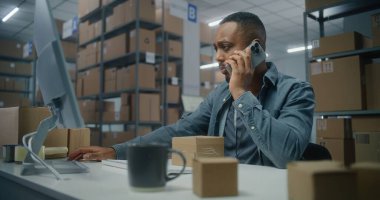 Sıralama merkezi: Afrikalı Amerikalı perakende satıcısı veya lojistik müdürü müşteriyle cep telefonuyla konuşur, teslimat için bilgisayarda paket bilgilerini kontrol eder. E-ticaret emriyle depo ya da depo..