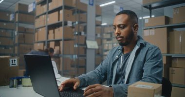 Posta servisi: Afro-Amerikan lojistik uzmanı barkod tarayıcılı paketleri tarıyor, müşteriye teslimata hazırlanıyor, dizüstü bilgisayar kullanıyor. Tasnif merkezi çalışanı kutu taşır. E-ticaret emirleri