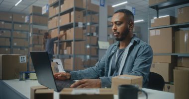 Posta servisi: Afro-Amerikan lojistik uzmanı barkod tarayıcılı paketleri tarıyor, müşteriye teslimata hazırlanıyor, dizüstü bilgisayar kullanıyor. Tasnif merkezi çalışanı kutu taşır. E-ticaret emirleri