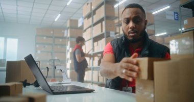 Afro-Amerikan postane çalışanlarının portresi barkod tarayıcısıyla tarama yapıyor, dizüstü bilgisayar kullanıyor, kameraya bakıyor. Teslimat için merkez çalışanları kutuları kontrol ediyor. Çevrimiçi mağaza deposu.