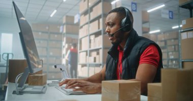 Afro-Amerikalı müşteri destek uzmanı, çağrı cihazını kullanarak, bilgisayar kullanarak, postanede ya da çevrimiçi depolamada çalışıyor. İşçiler karton kutulara bakıyor..