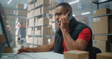 Postane veya E-ticaret mağazası depolama tesisi: Cep telefonuyla konuşan Afrikalı Amerikalı lojistik koordinatörü, bilgisayar kullanıyor. Merkez çalışanları nakliye için karton kutuları kontrol ediyor.