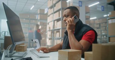 E-ticaret veya posta servisinin depo perakende tesisi: Afrikalı Amerikalı lojistik koordinatörü telefonla konuşuyor, bilgisayar kullanıyor. Teslimat için karton kutuları kontrol eden sıralama merkezi çalışanları.