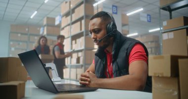 Kulaklıkla Afrikalı Amerikalı lojistik koordinatörünün portresi müşteriyle konuşuyor, posta servisinde ya da çevrimiçi depolamada dizüstü bilgisayarla çalışıyor, kameraya bakıyor. Sıralama merkezi çalışanları kutucukları işaretliyor