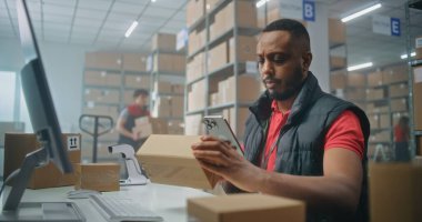 Afro-Amerikan lojistik müdürü akıllı telefon kullanarak paketleri tarıyor, posta servisi deposunda çalışıyor. Nakliye için karton kutular taşıyan tasnif merkezi çalışanları. E-ticaret emirleriyle birlikte depolama.