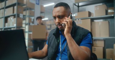 E-ticaret mağazası veya postanenin depo perakende satış tesisi: Afrikalı Amerikalı lojistik koordinatörü telefonla konuşuyor, dizüstü bilgisayar kullanıyor. Teslimat için karton kutular taşıyan tasnif merkezi çalışanları.
